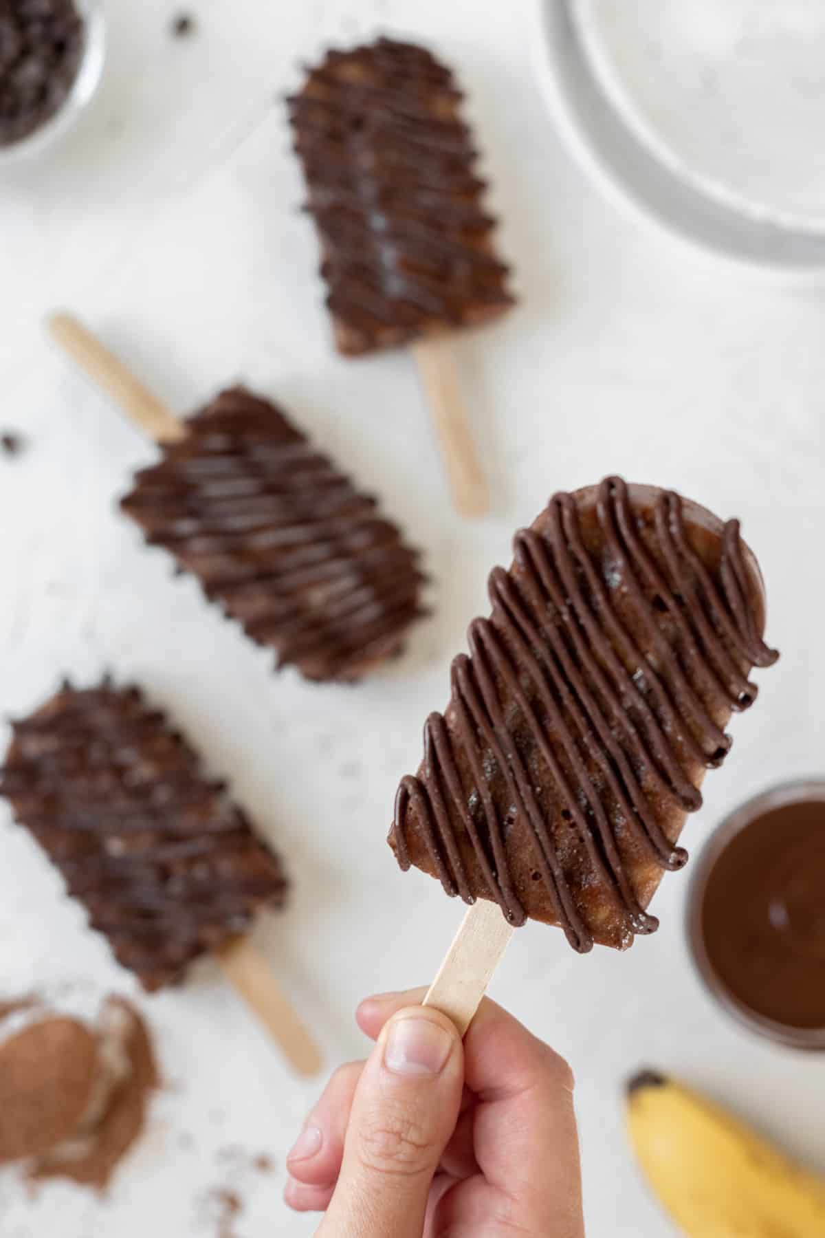 A hand holding a banana fudge popsicle in the air with 3 other popsicles on a white background below it.