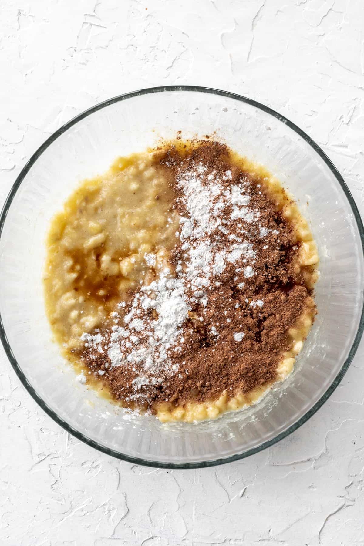 Mashed bananas in a glass bowl with cacao powder, corn starch, and vanilla sprinkled on top.