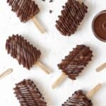 Creamy banana fudge chocolate popsicles spread out on a white background with melted chocolate and chocolate chops around them.