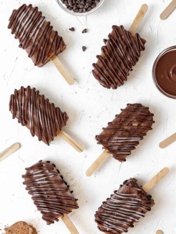 Creamy banana fudge chocolate popsicles spread out on a white background with melted chocolate and chocolate chops around them.