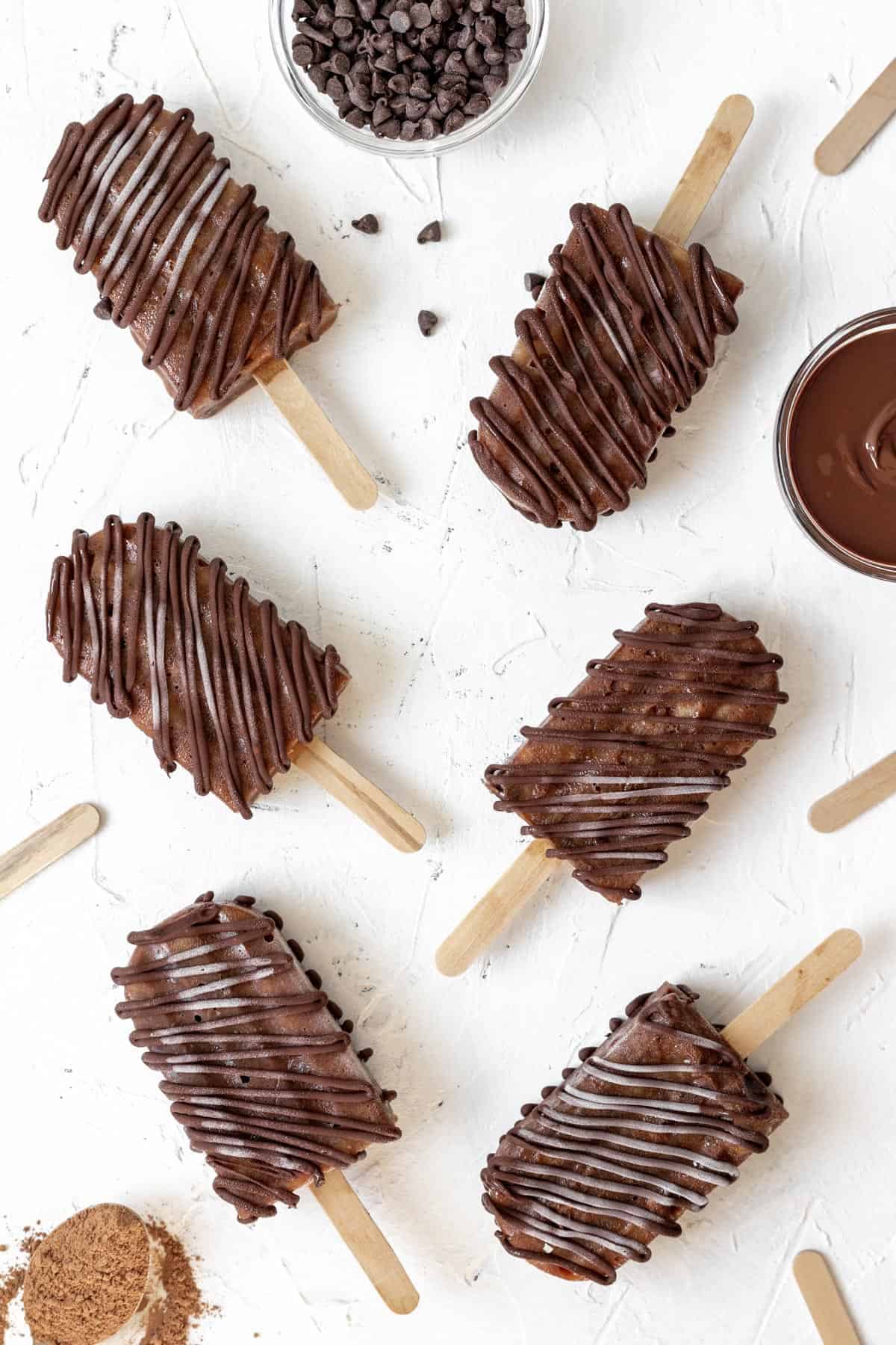 Creamy banana fudge chocolate popsicles spread out on a white background with melted chocolate and chocolate chops around them.