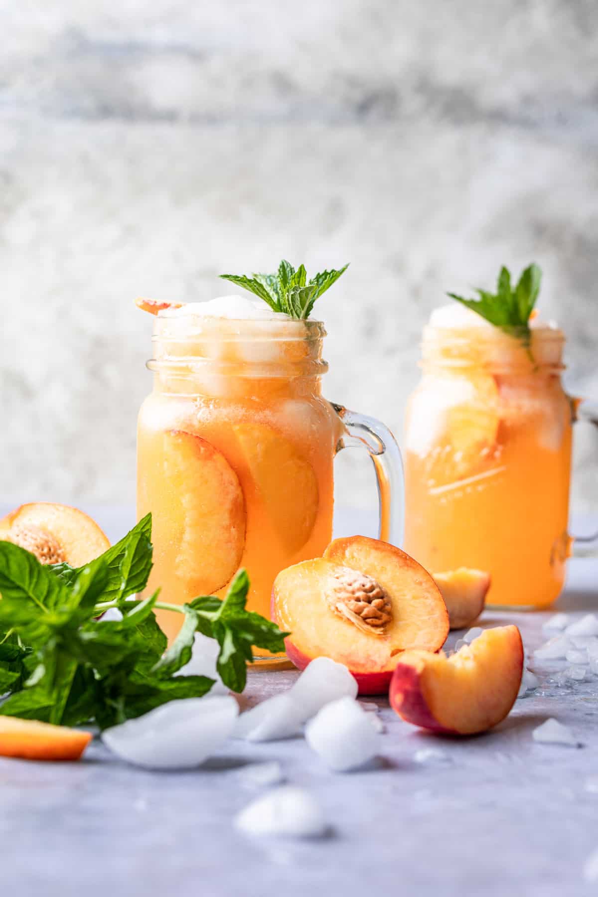 Two bourbon peach smashes with ice, peaches, and mint around them.