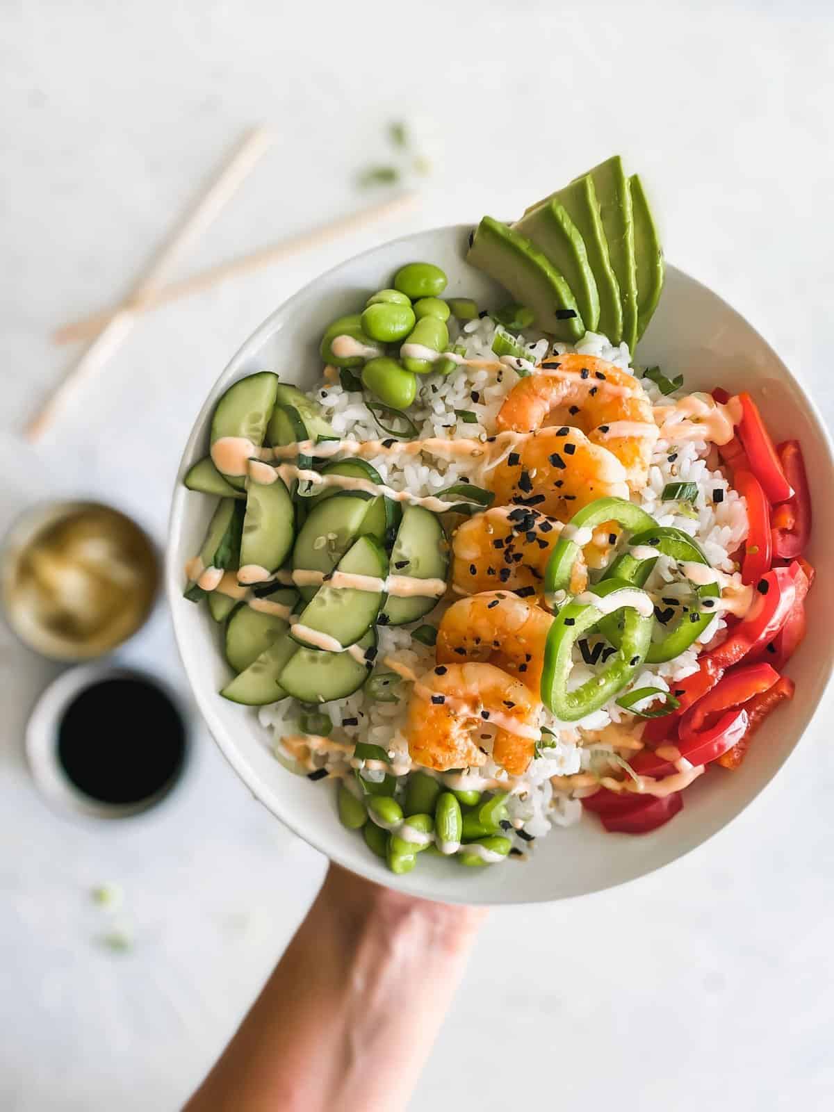 shrimp poke bowl raised in the air with chopsticks, sushi ginger, and soy sauce in background