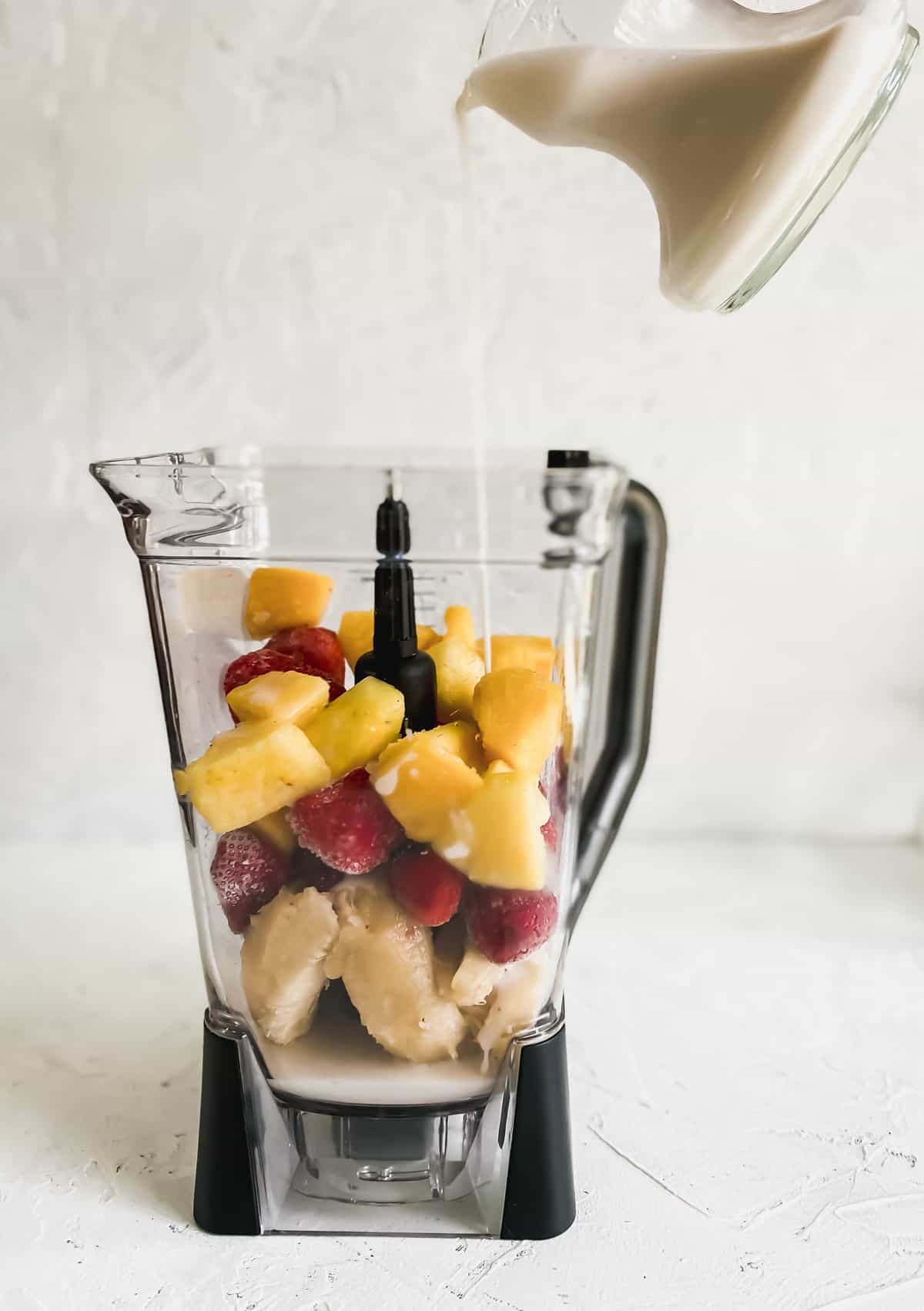 pouring almond milk into a blender with frozen pineapples, strawberries, and bananas