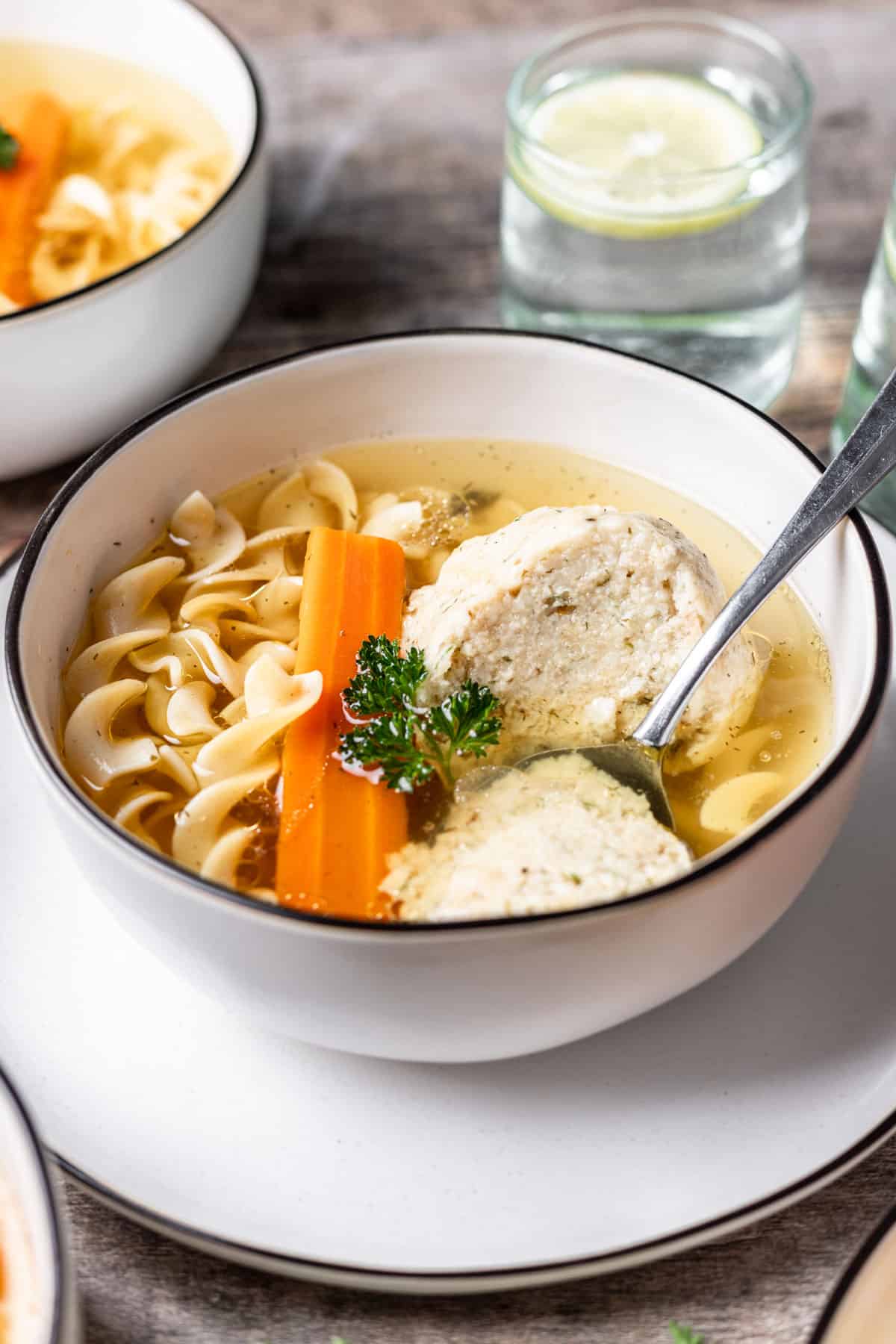 Instant pot matzo ball soup in a white bowl with a spoon and a glass of water next to it.