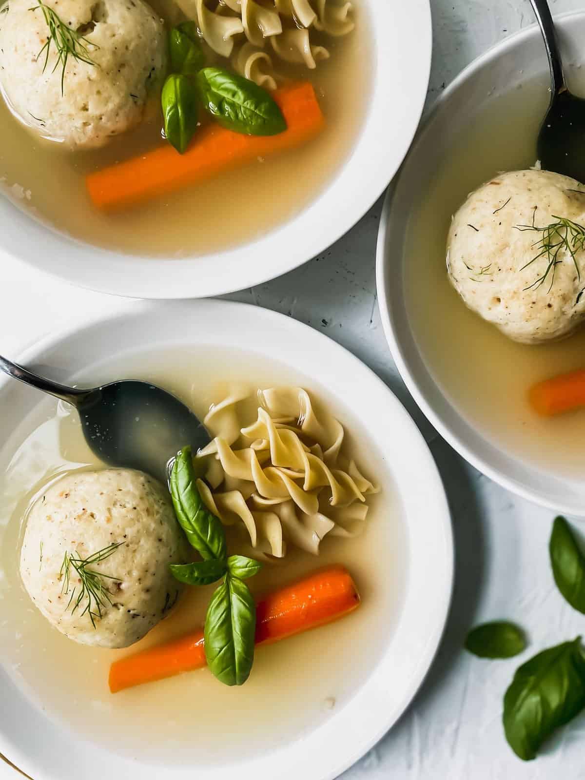 3 bowls of matzo ball soup in white bowls with spoons, carrots, and noodles