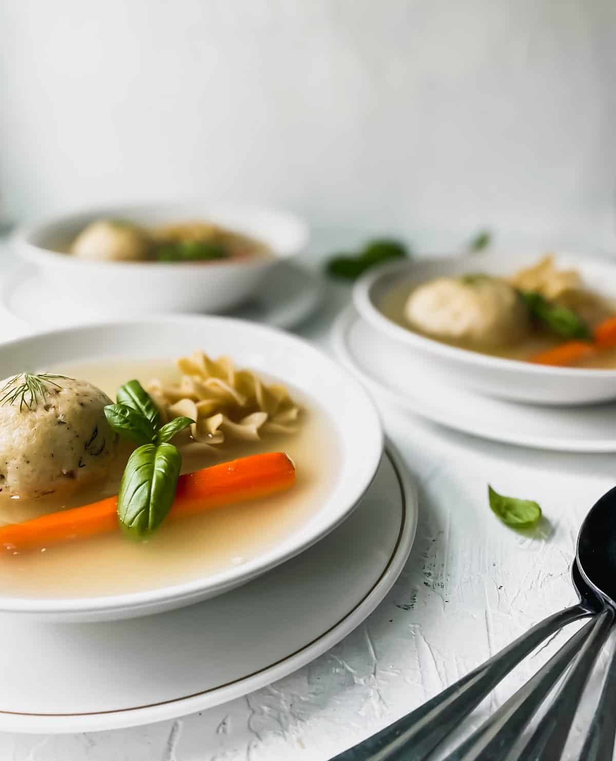 3 bowls of matzo ball soup with metal spoons on the side
