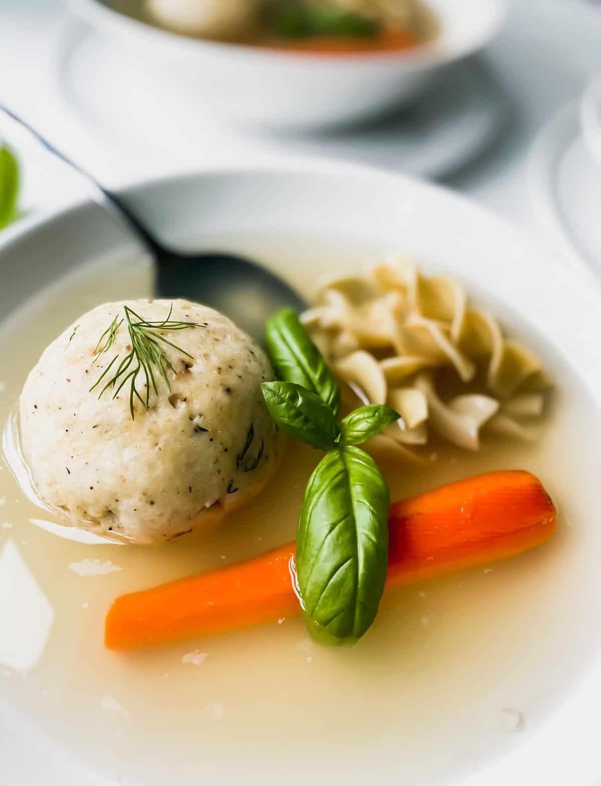 matzo ball soup in a white bowl with a spoon, noodles, and a carrot