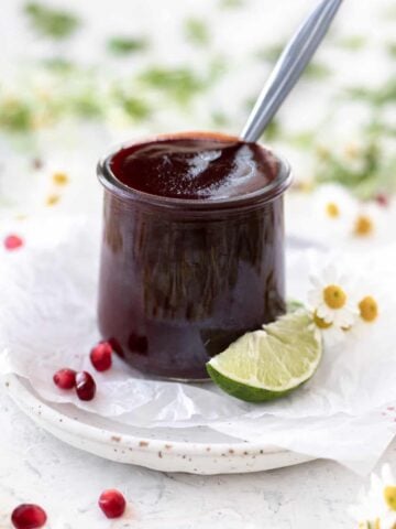 Pomegranate bbq sauce in a clear jar with a spoon sticking out and limes, pomegranate seeds, and flowers around it.