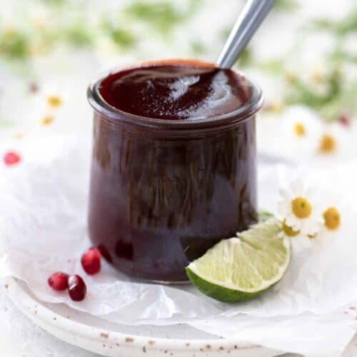 Pomegranate bbq sauce in a clear jar with a spoon sticking out and limes, pomegranate seeds, and flowers around it.