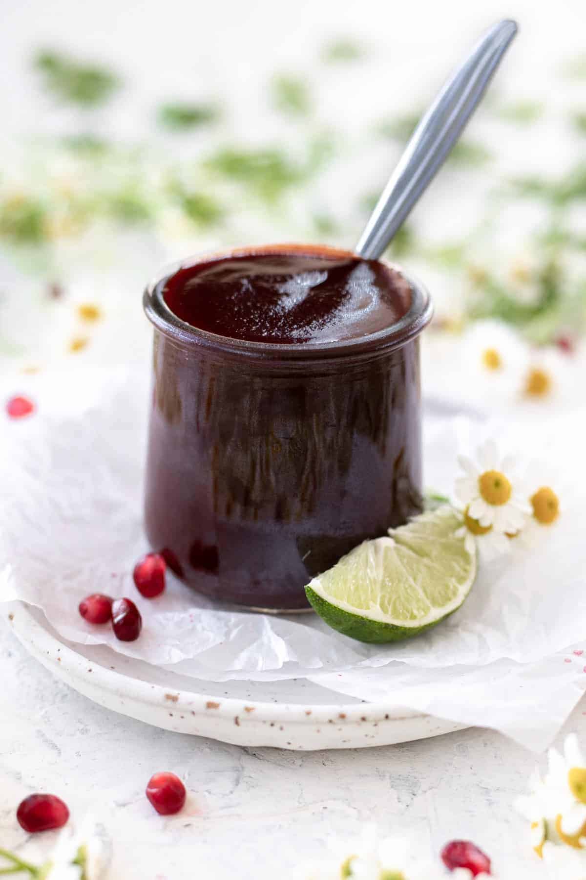 Pomegranate bbq sauce in a clear jar with a spoon sticking out and limes, pomegranate seeds, and flowers around it.