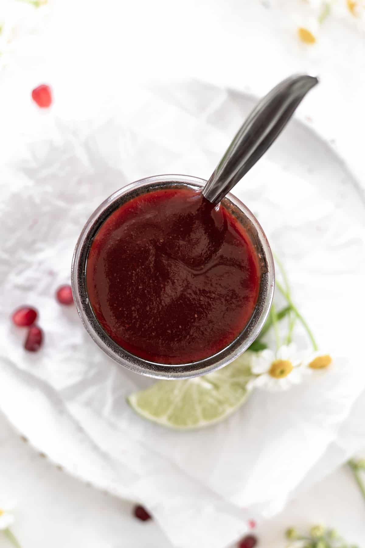 A birds eye view of pomegranate bbq sauce in a clear jar with a spoon sticking out.