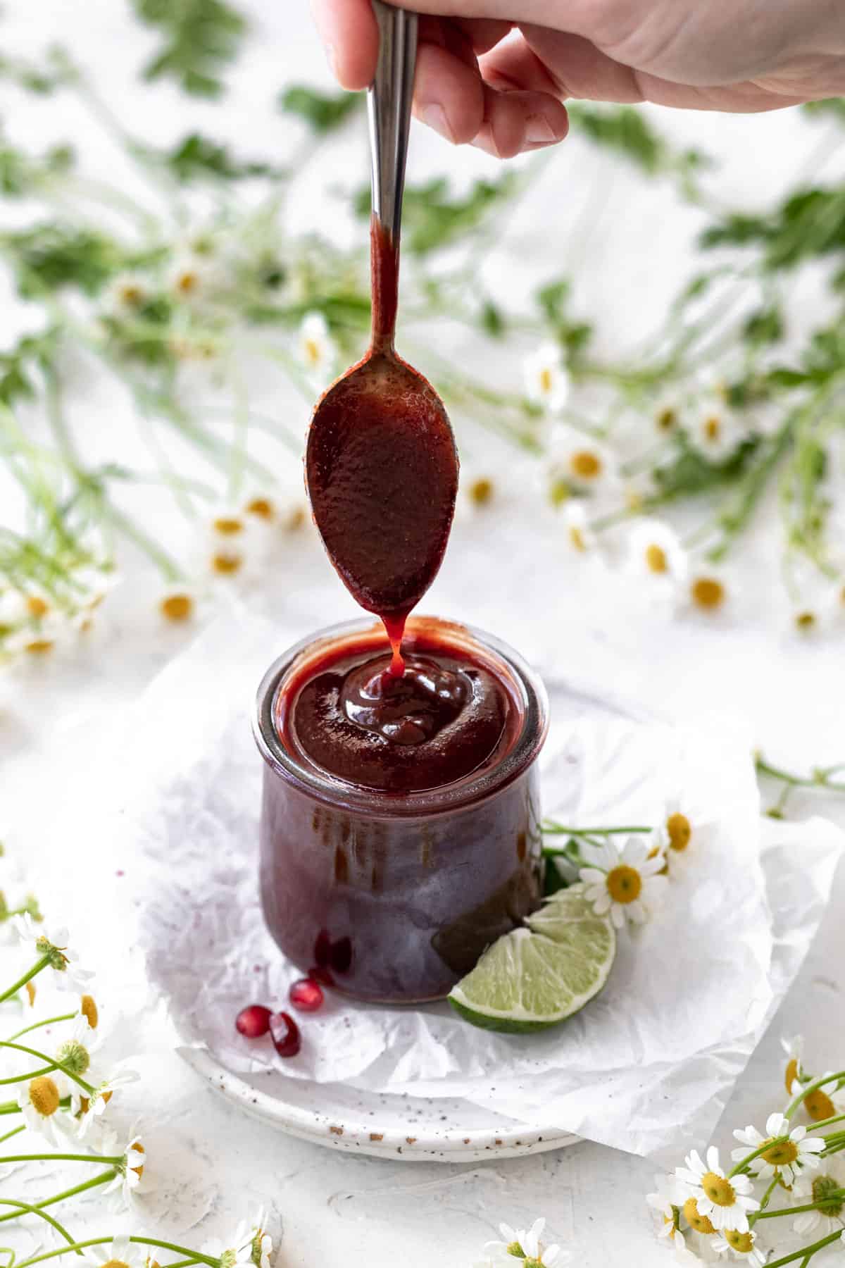 A hand drizzling sweet and tangy sauce into a glass jar surrounded by pomegranate seeds, lime, and flowers.
