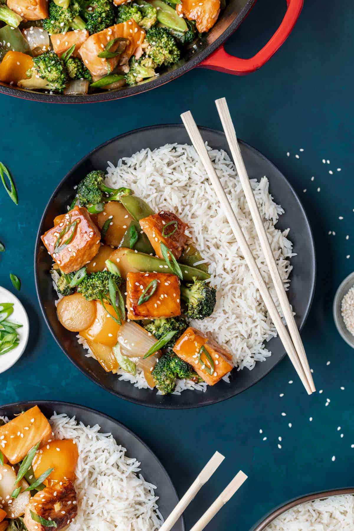 Teriyaki salmon stir fry on a bed of rice in a black bowl with chopsticks.