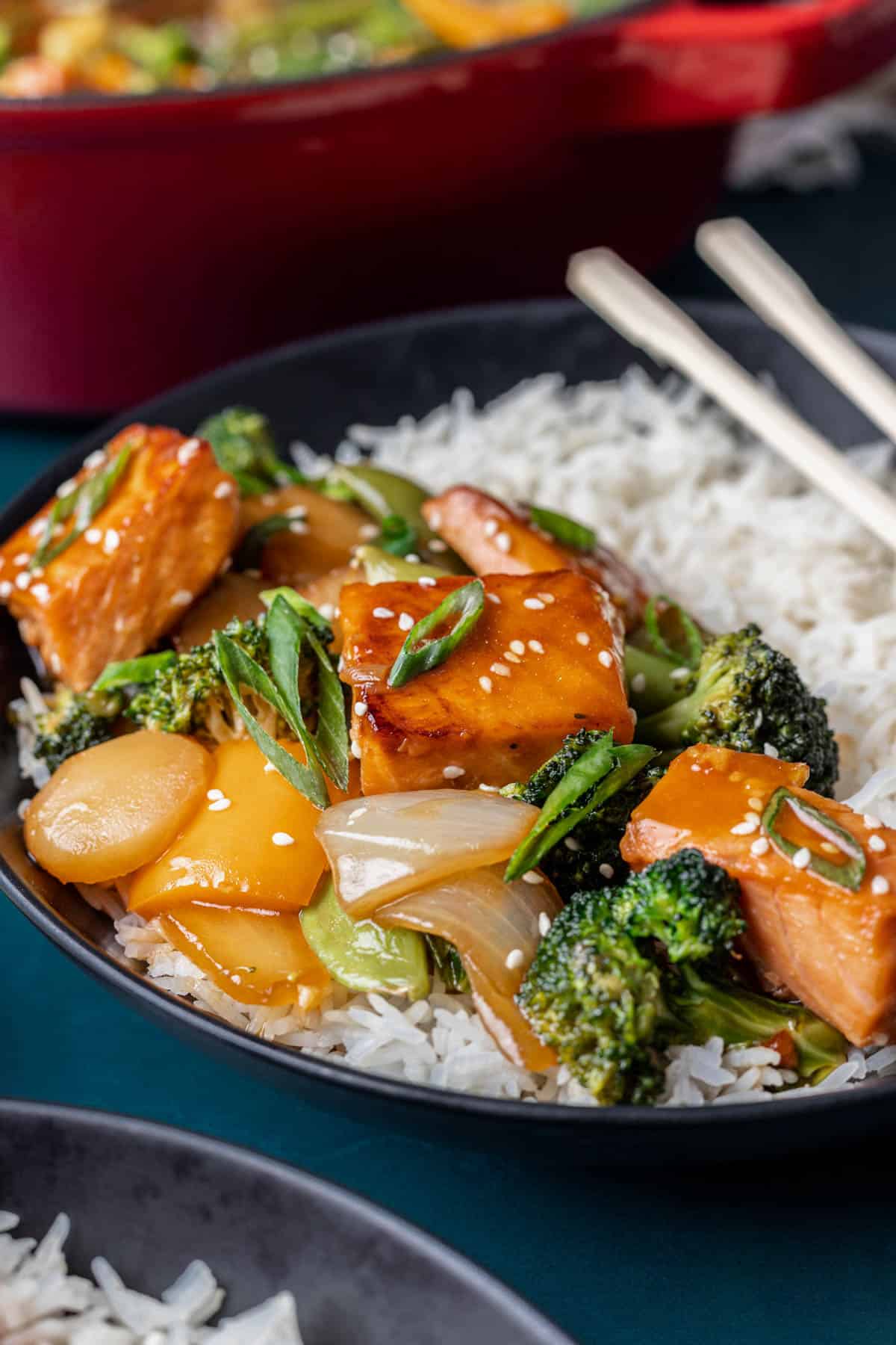 A piece of salmon glazed in teriyaki sauce on top of stir fried vegetables and white rice.