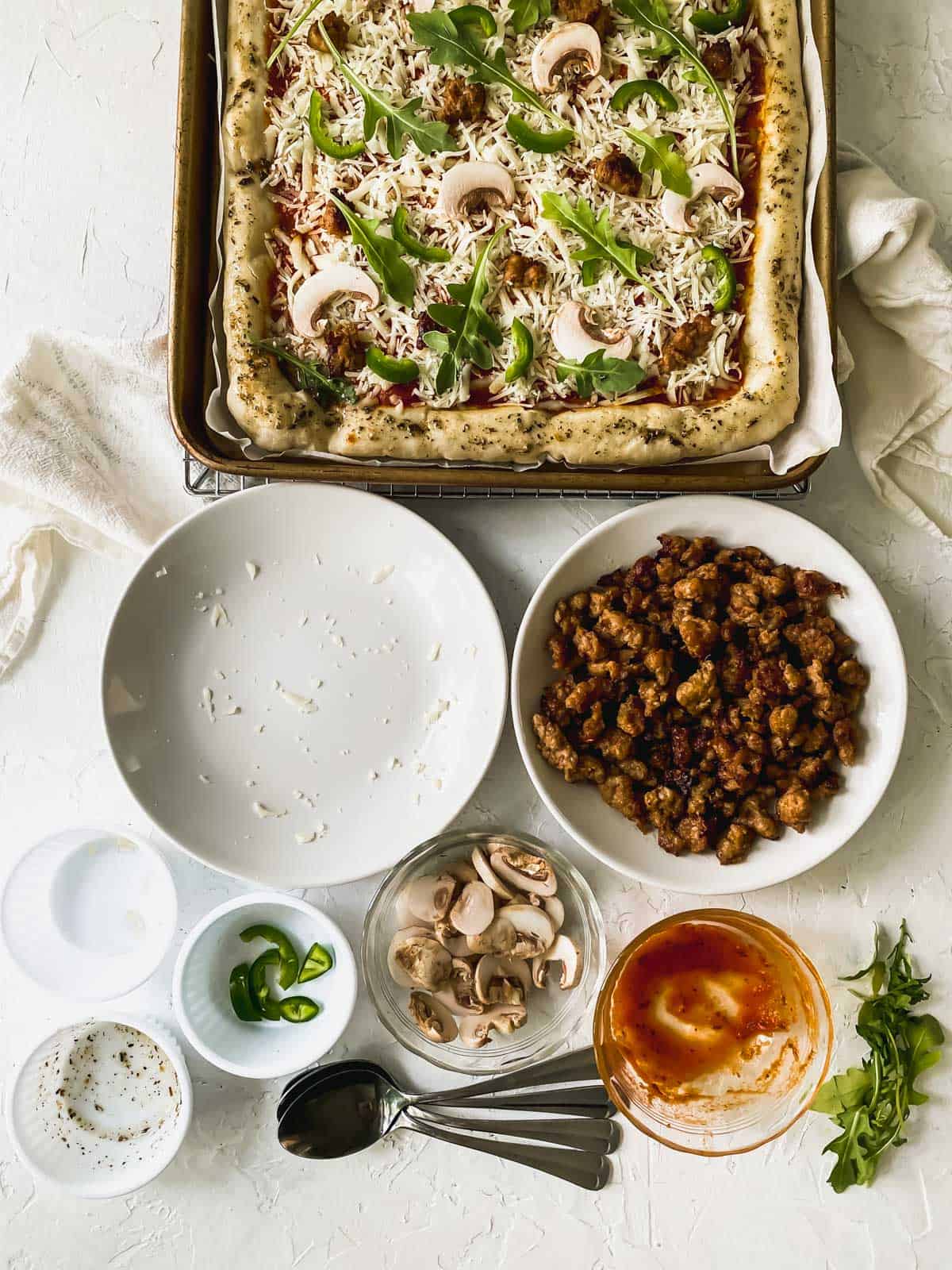 sauce, cheese, and toppings on top of pizza dough with bowls around it