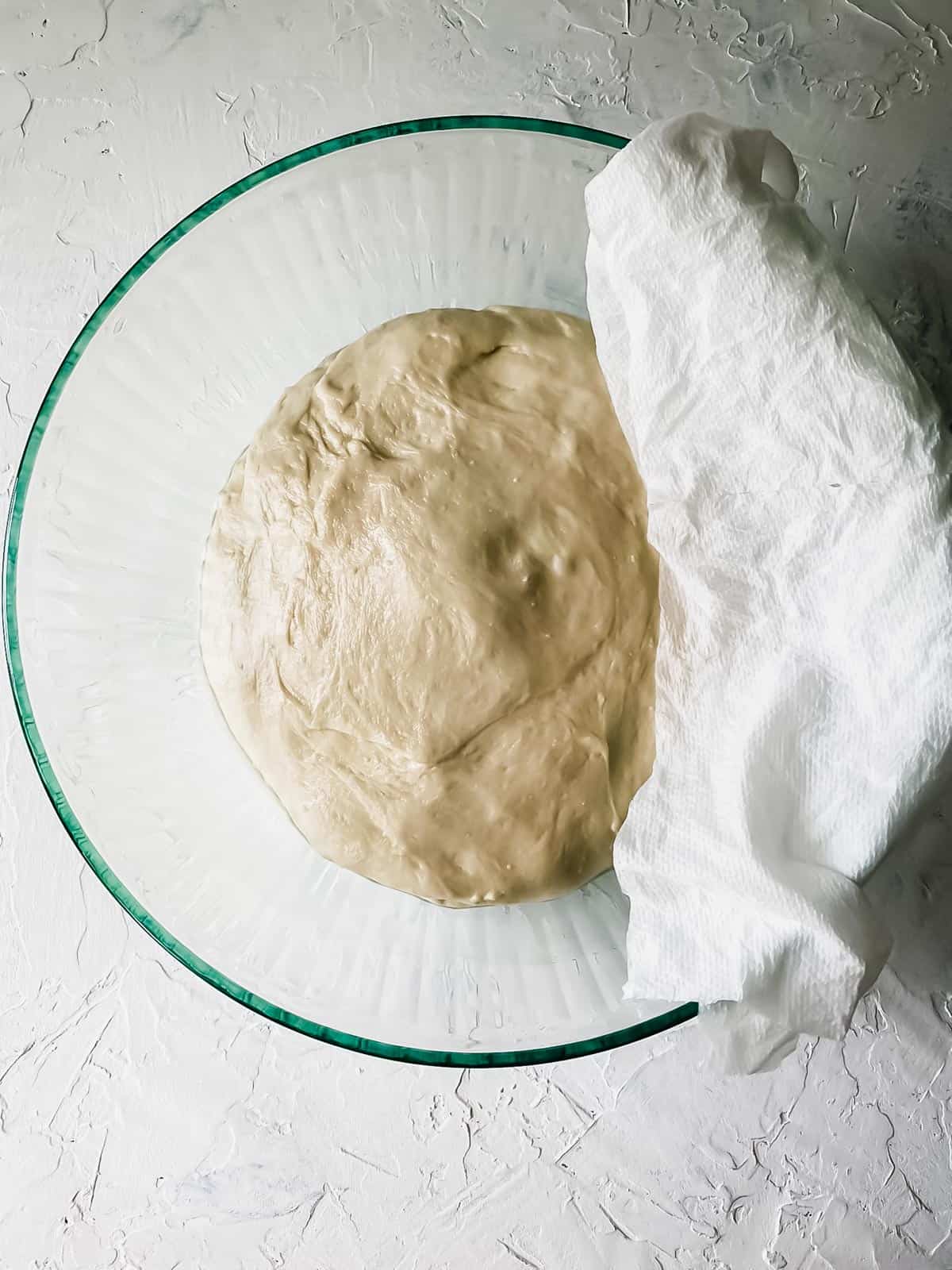 pizza dough in a glass bowl with a damp paper towel on the side