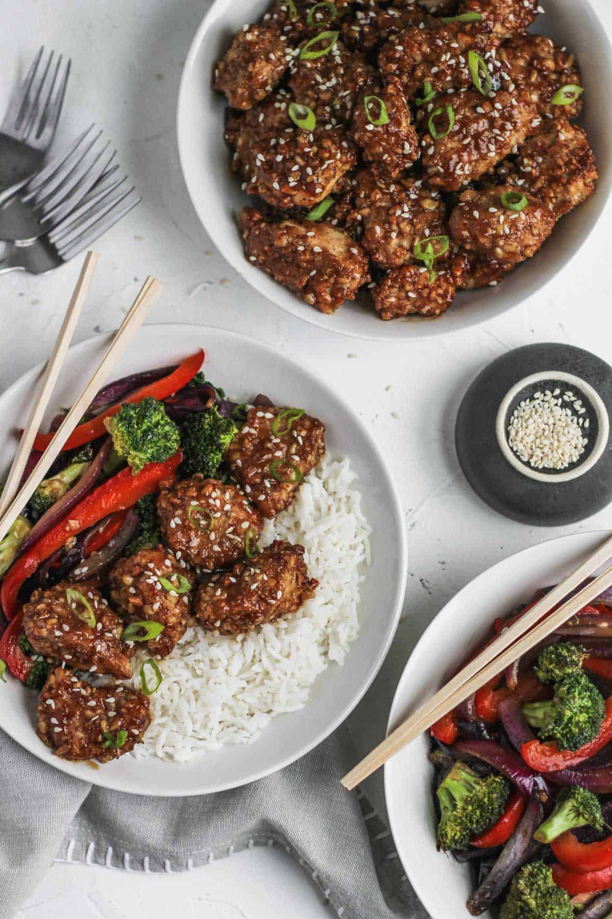 Air fryer almond chicken in white bowls with a side of sesame seeds and stir fried veggies.