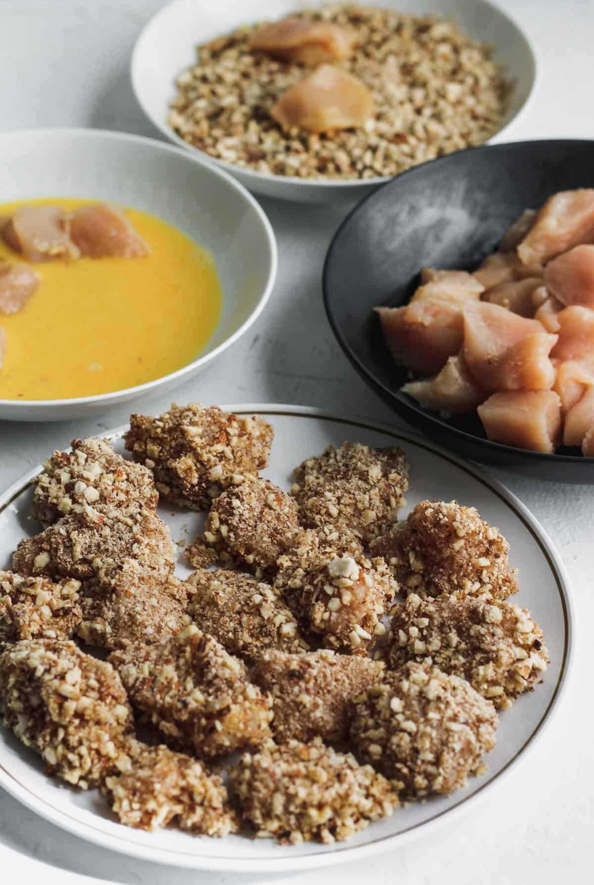 Separate bowls with the beaten egg, crushed almonds, raw chicken, and almond coated chicken. 