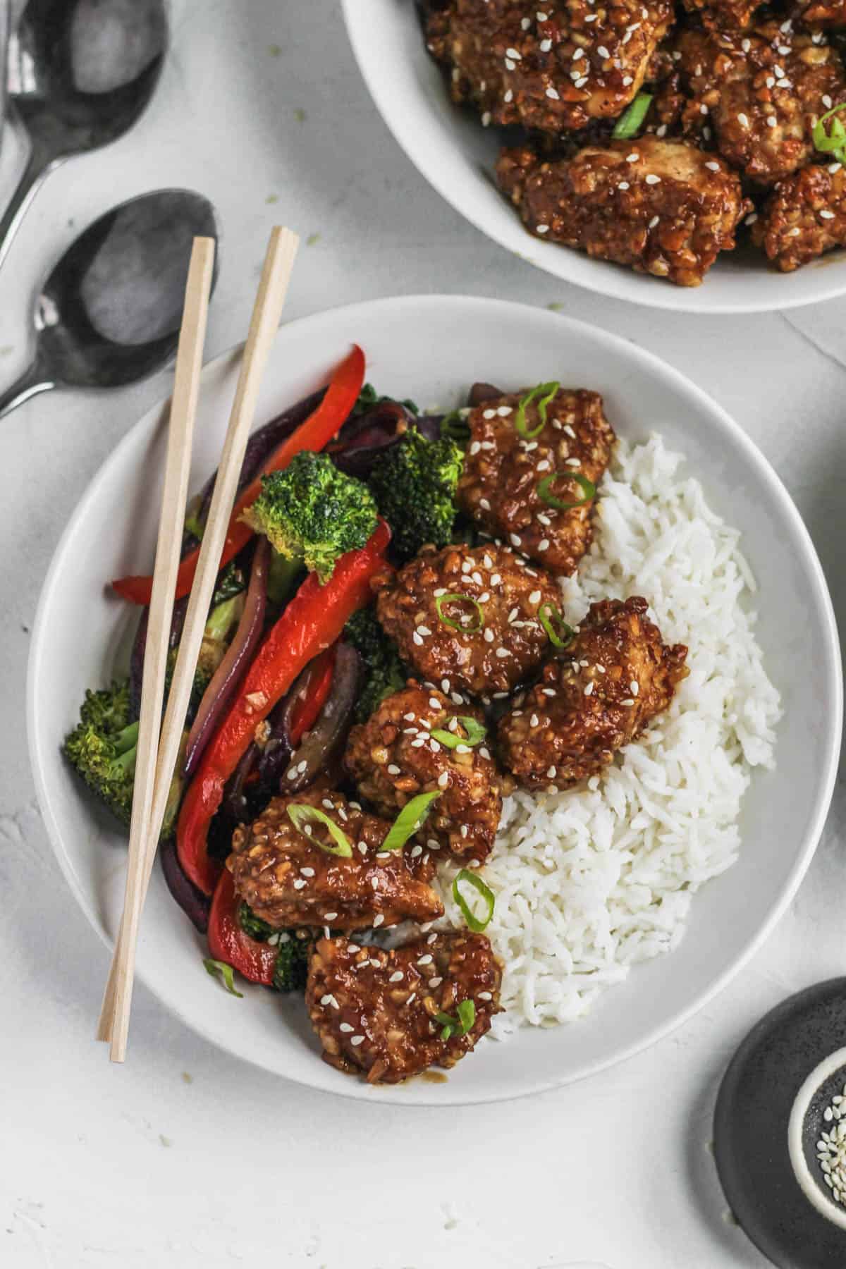 Crispy chicken in honey soy sauce in a bowl with stir fried veggies and white rice with chopsticks and sesame seeds on the side.