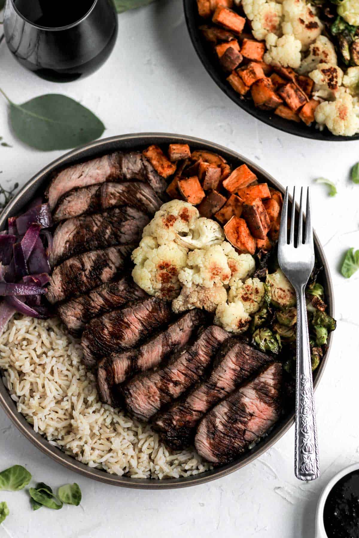 Glazed balsamic steak and veggie bowls with red wine, a fork, and brussels sprout leaves around it.