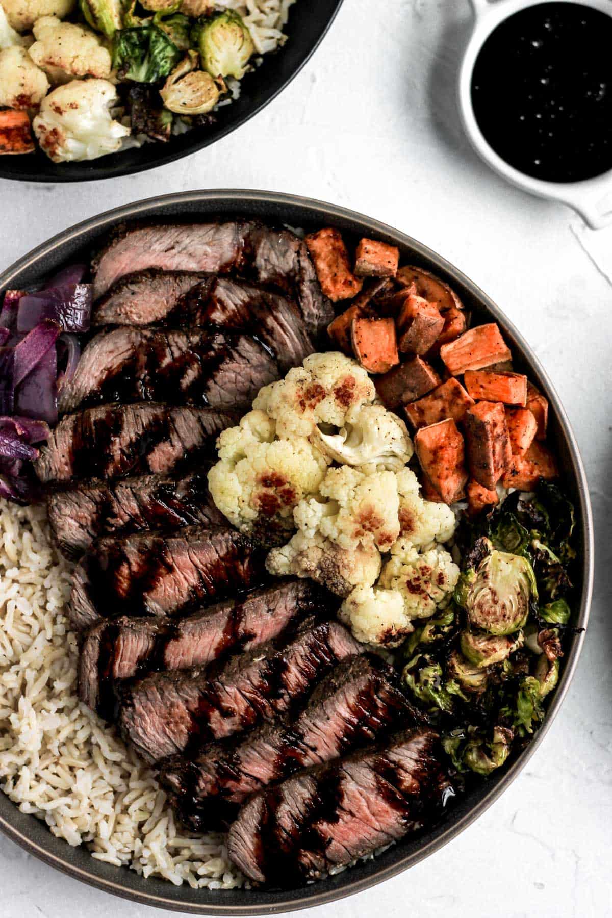 Glazed balsamic steak and veggies over brown rice in a dark gray bowl with a side of balsamic reduction. 