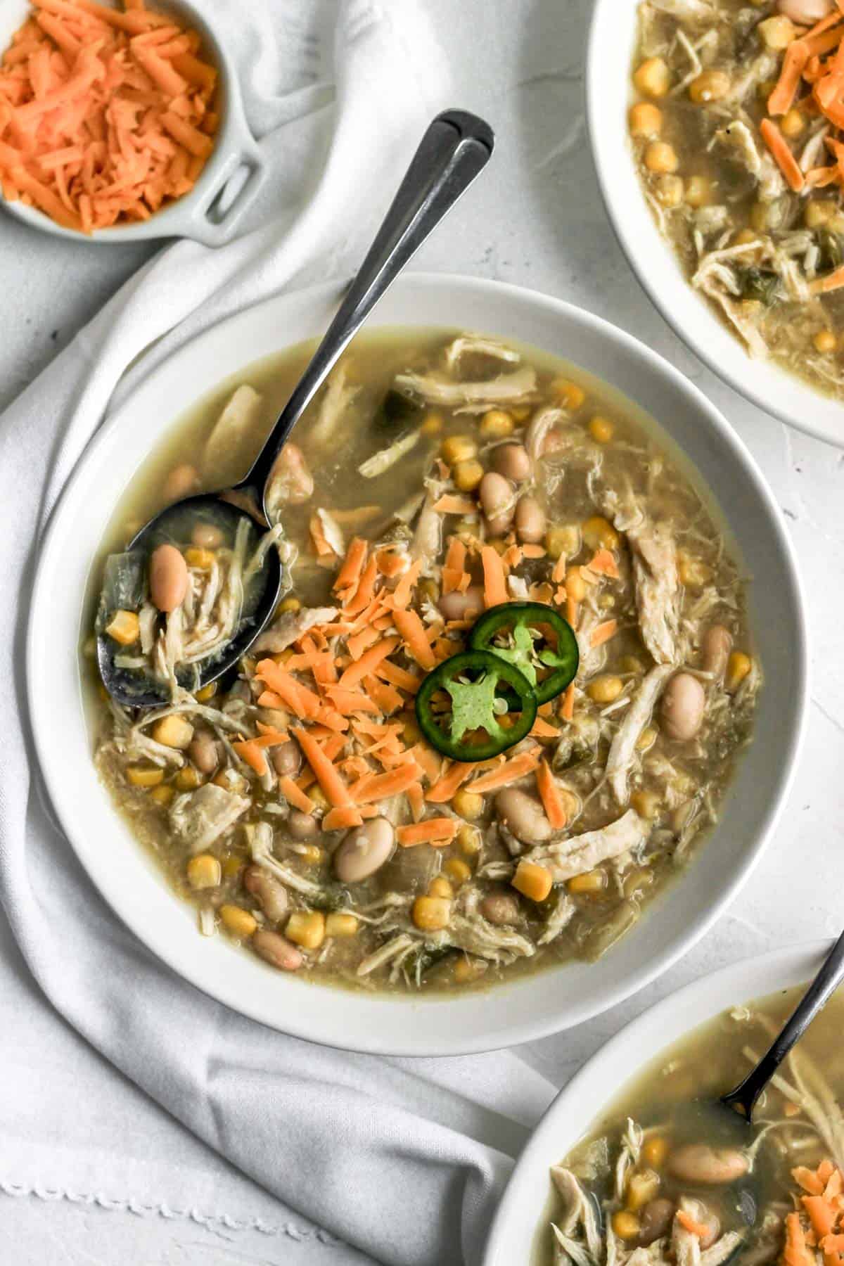 White chicken chili in 3 white bowls with a side of cheddar cheese and a white napkin.