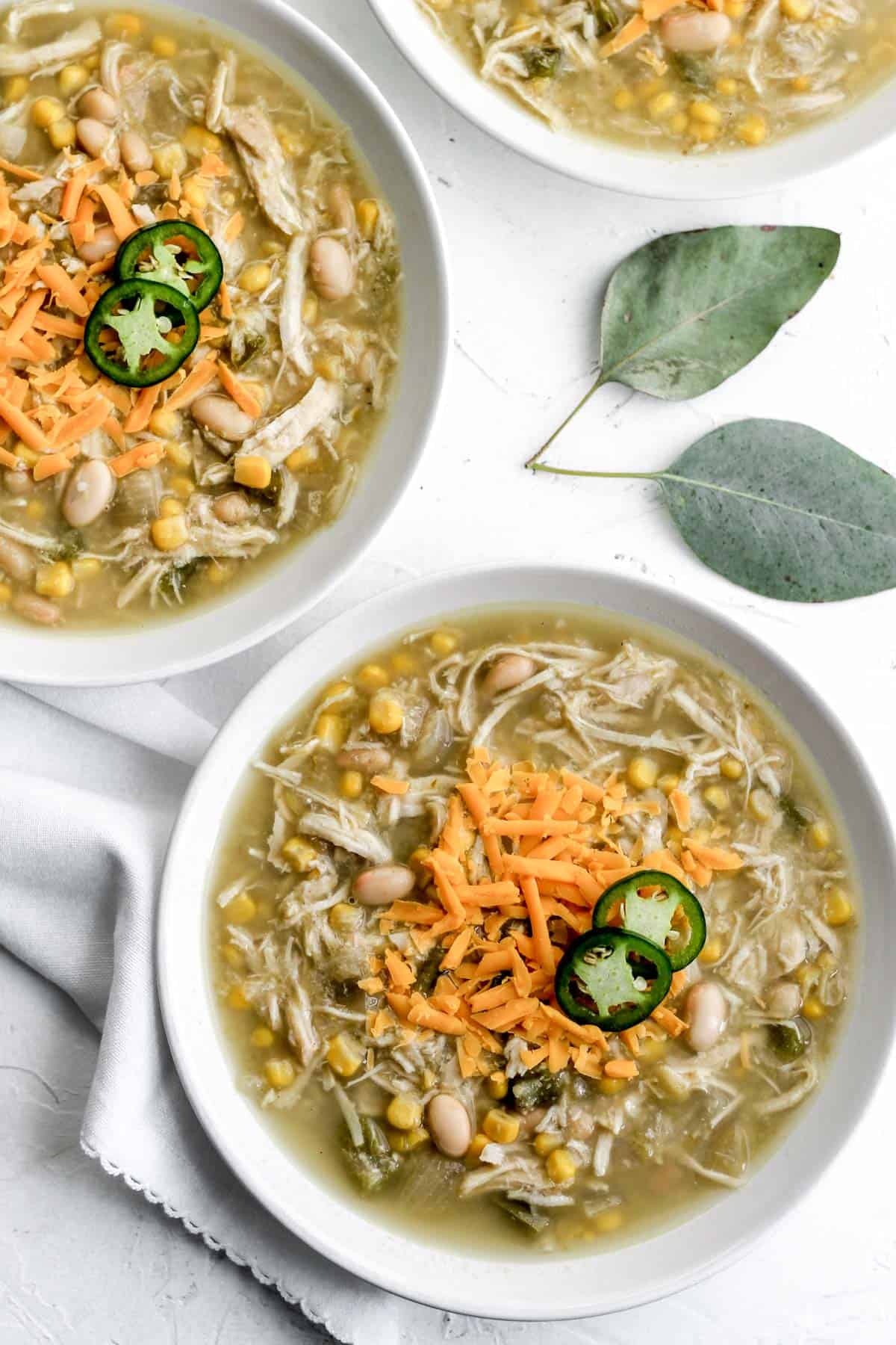 White chicken chili in 2 white bowls with a white napkin and 2 eucalyptus leaves next to them.