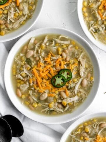 Healthy chicken chili in a white bowl with silver spoons and 3 additional bowls surrounding it.