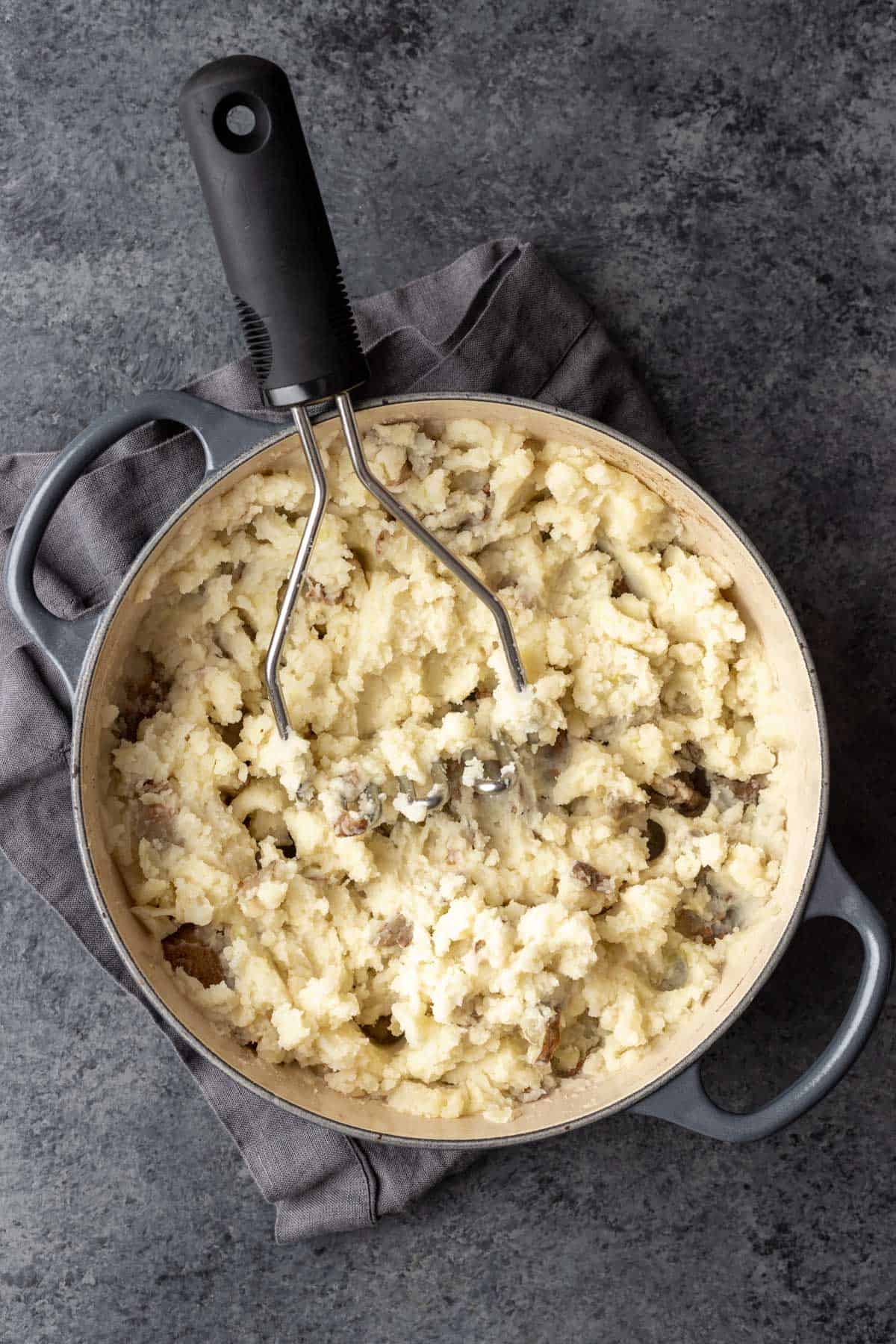 Mashed potatoes in a large pot with a potato masher.