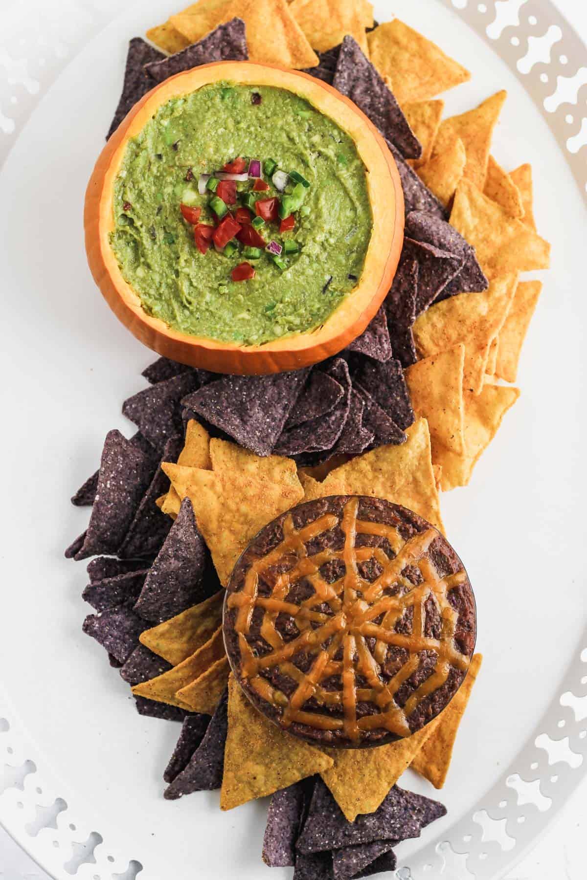 Guacamole and black bean dip in pumpkins on a white platter with a swirl of tortilla chips between.