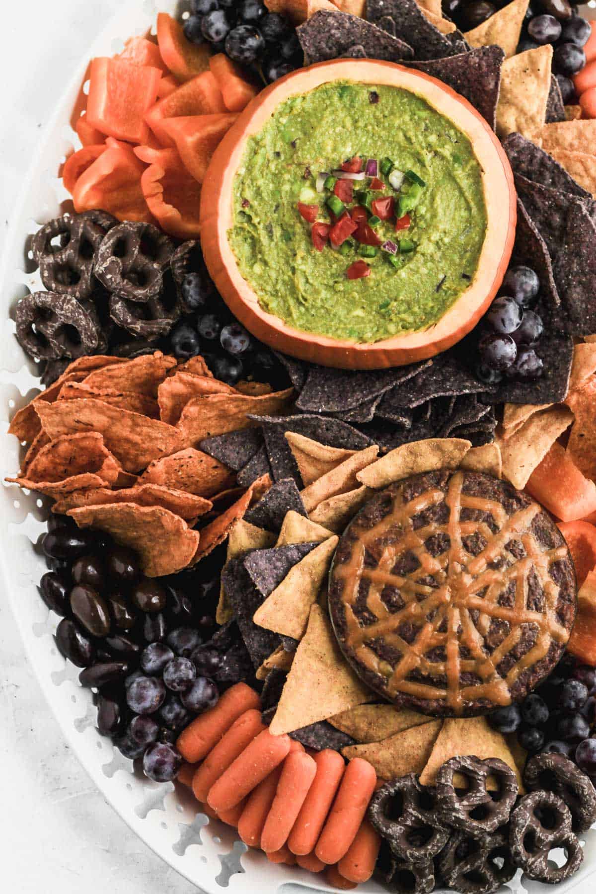 Halloween inspired guacamole snack board with dips, chips, chocolate almonds, carrots, peppers, chocolate pretzels, chocolate almonds, and grapes on a white platter.