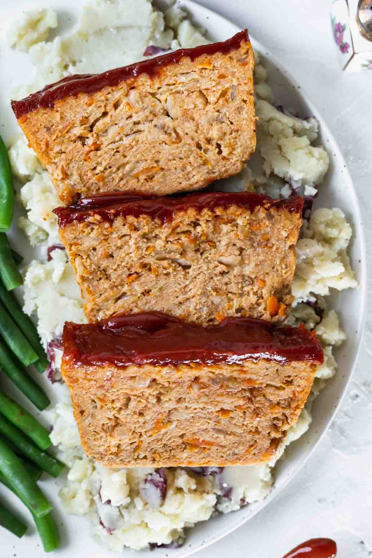 A closeup view of ground chicken meatloaf on a dinner plate with green beans and mashed potatoes.