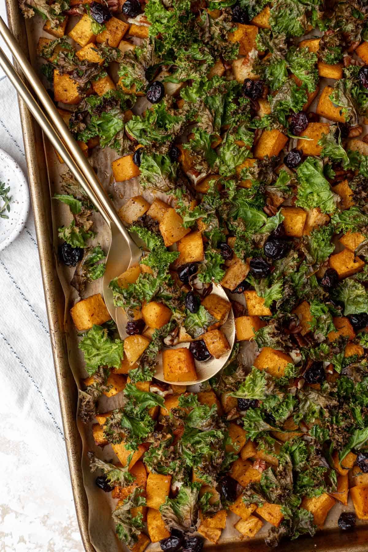 Roasted maple cinnamon butternut squash, crispy kale, baked apples, and dried cranberries on a sheet pan with two serving spoons for scooping.