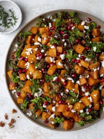 Roasted butternut squash with baked apples and crispy kale in a serving bowl with pomegranate seeds and feta on top.