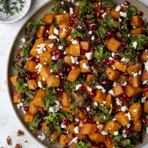 Roasted butternut squash with baked apples and crispy kale in a serving bowl with pomegranate seeds and feta on top.