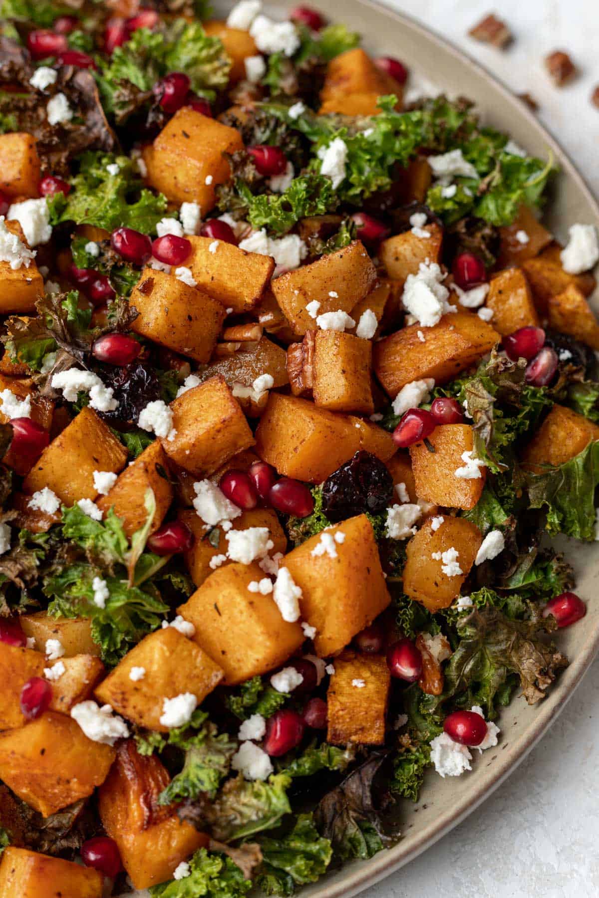 A closeup view of maple cinnamon squash, baked apples, kale chips, feta, pomegranate seeds, and toasted pecans in a beige serving bowl.