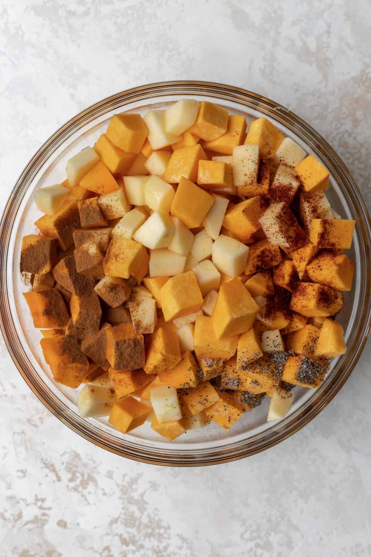 Diced and peeled butternut squash and apples in a glass mixing bowl with seasonings on top.