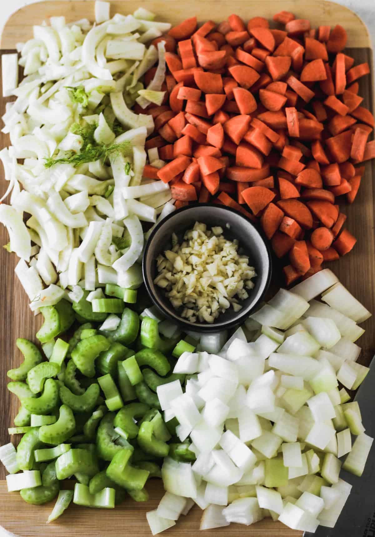 Chopped onions, sliced fennel, diced carrots and celery, and minced garlic on a wooden cutting board.