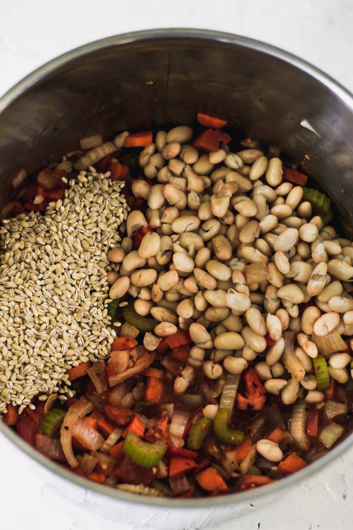 Vegetables cooking in a tall pot topped with great northern beans and dry barley.