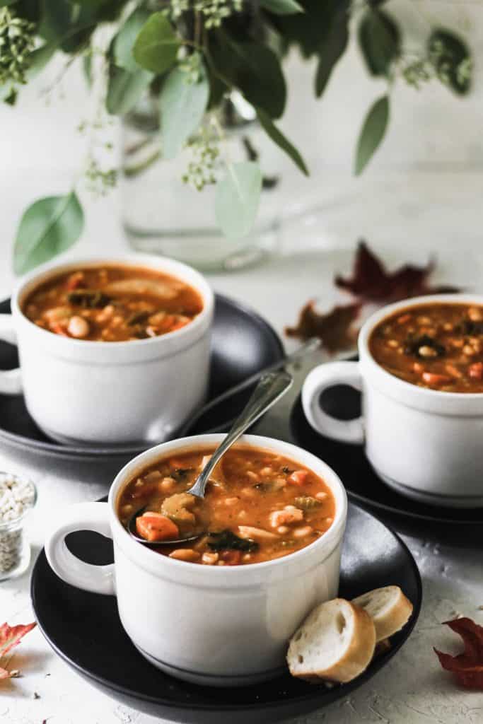One pot vegetable bean and barley soup in 3 white mugs on top of black plates with metal spoons.