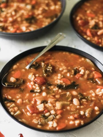 One pot vegetable, bean, and barley soup in 3 black bowls with a silver spoon.