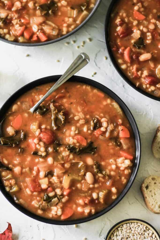 Vegetable, bean, and barley soup in a black bowl with a spoon and side of crusty bread.