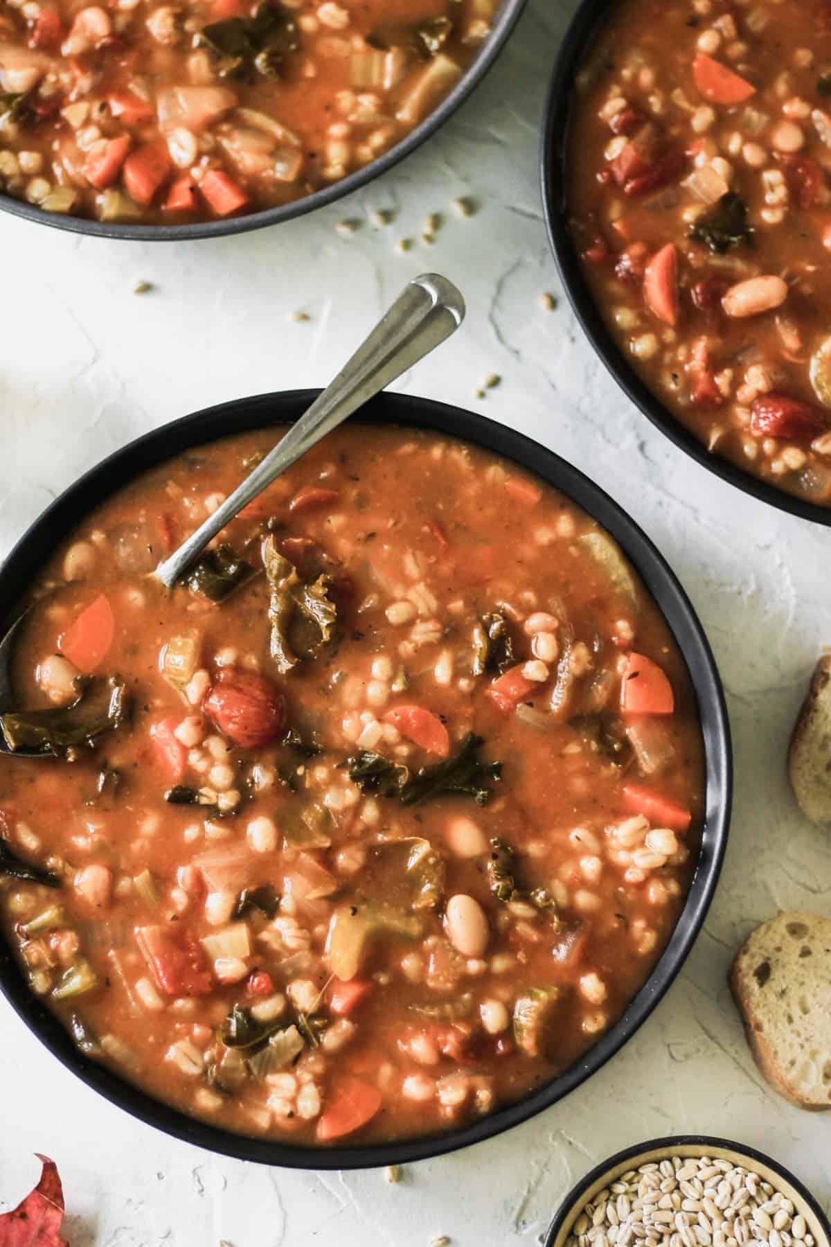 Vegetable, bean, and barley soup in a black bowl with a spoon and side of crusty bread.