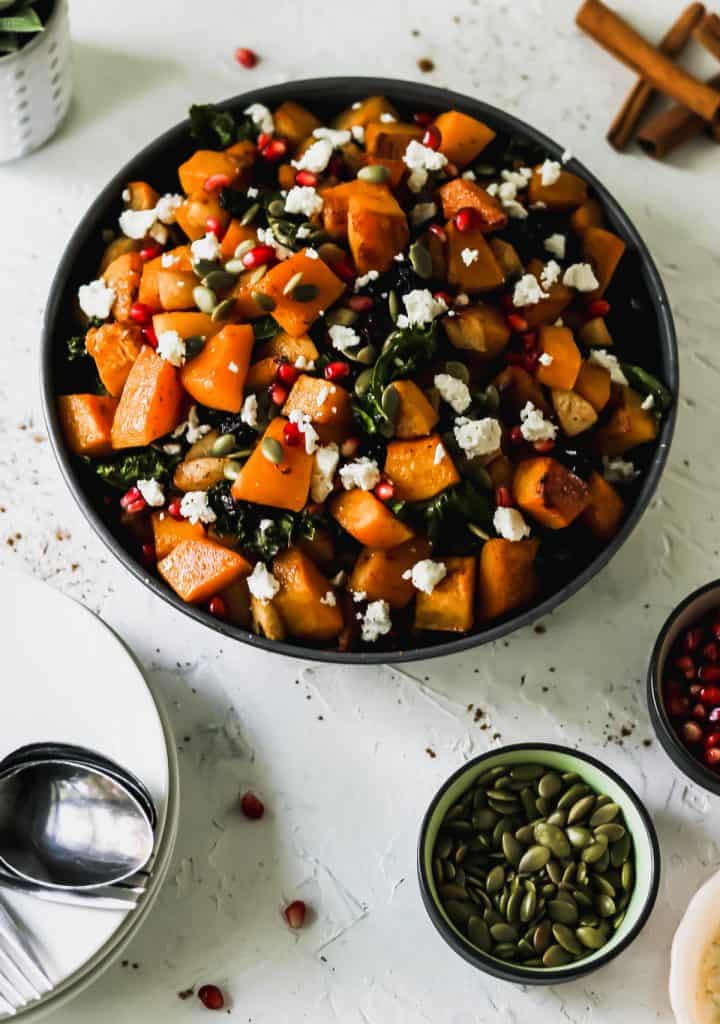 Roasted squash, apples, kale, and cranberries in a black serving bowl with white plates, a serving spoon, cinnamon sticks, pumpkin seeds, and pomegranate seeds around it.