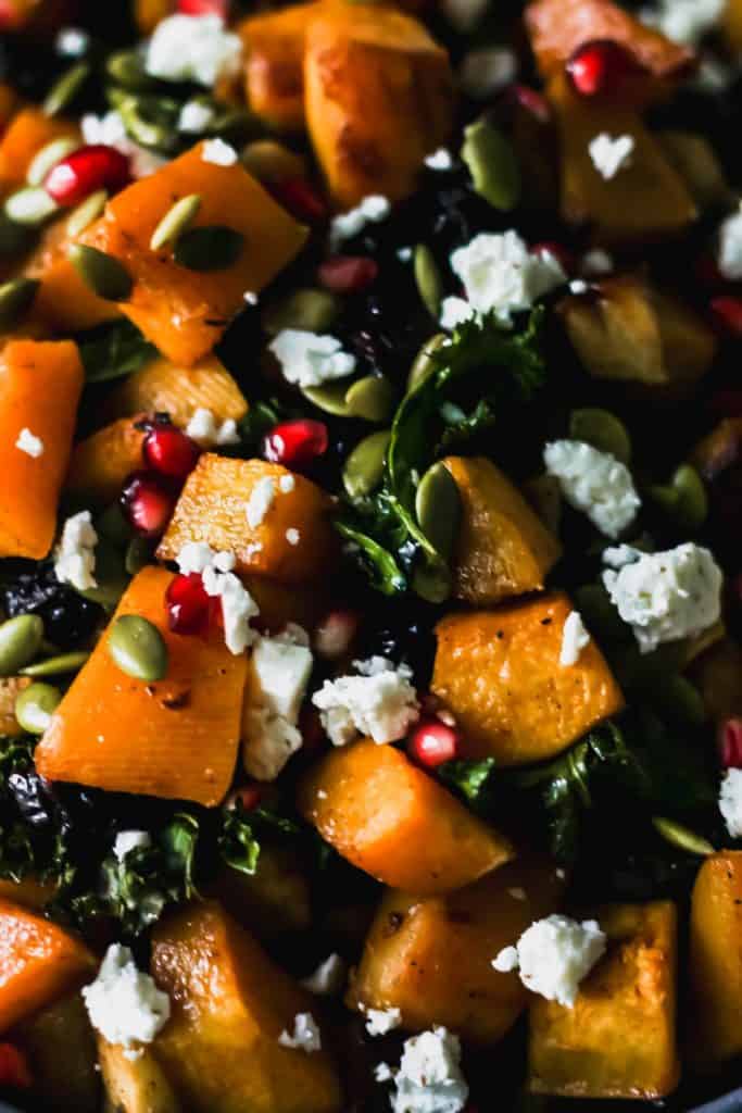 Close up of roasted butternut squash with apples and kale topped with pumpkin seeds, feta, and pomegranate seeds.