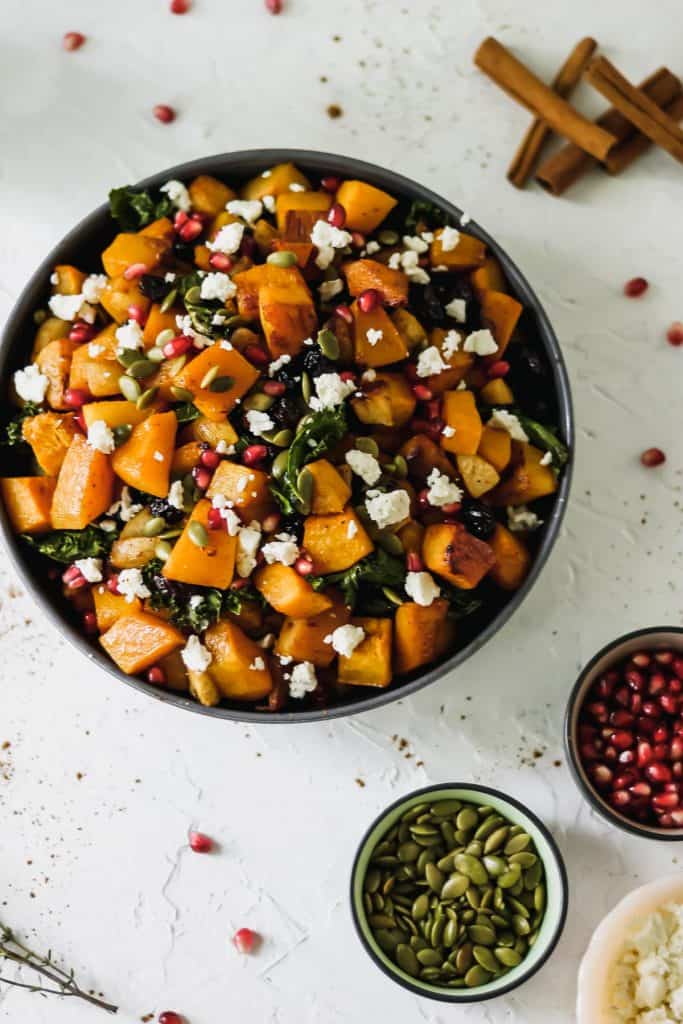 Roasted butternut squash with apples and kale in a black bowl with pumpkin seeds, pomegranate seeds, feta, thyme, and cinnamon sticks around it.