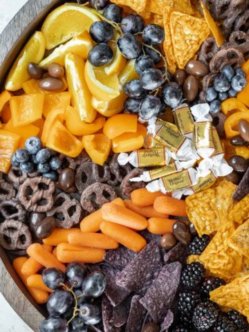 Spooky Halloween snack board with orange slices, caramels, carrots, nacho cheese chips, blue tortilla chips, chocolate pretzels, grapes, and blueberries.