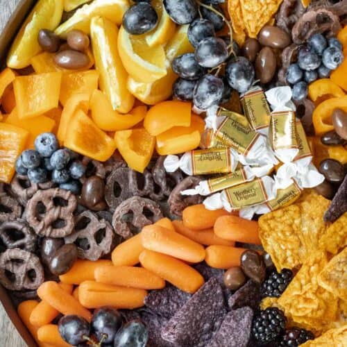 Spooky Halloween snack board with orange slices, caramels, carrots, nacho cheese chips, blue tortilla chips, chocolate pretzels, grapes, and blueberries.