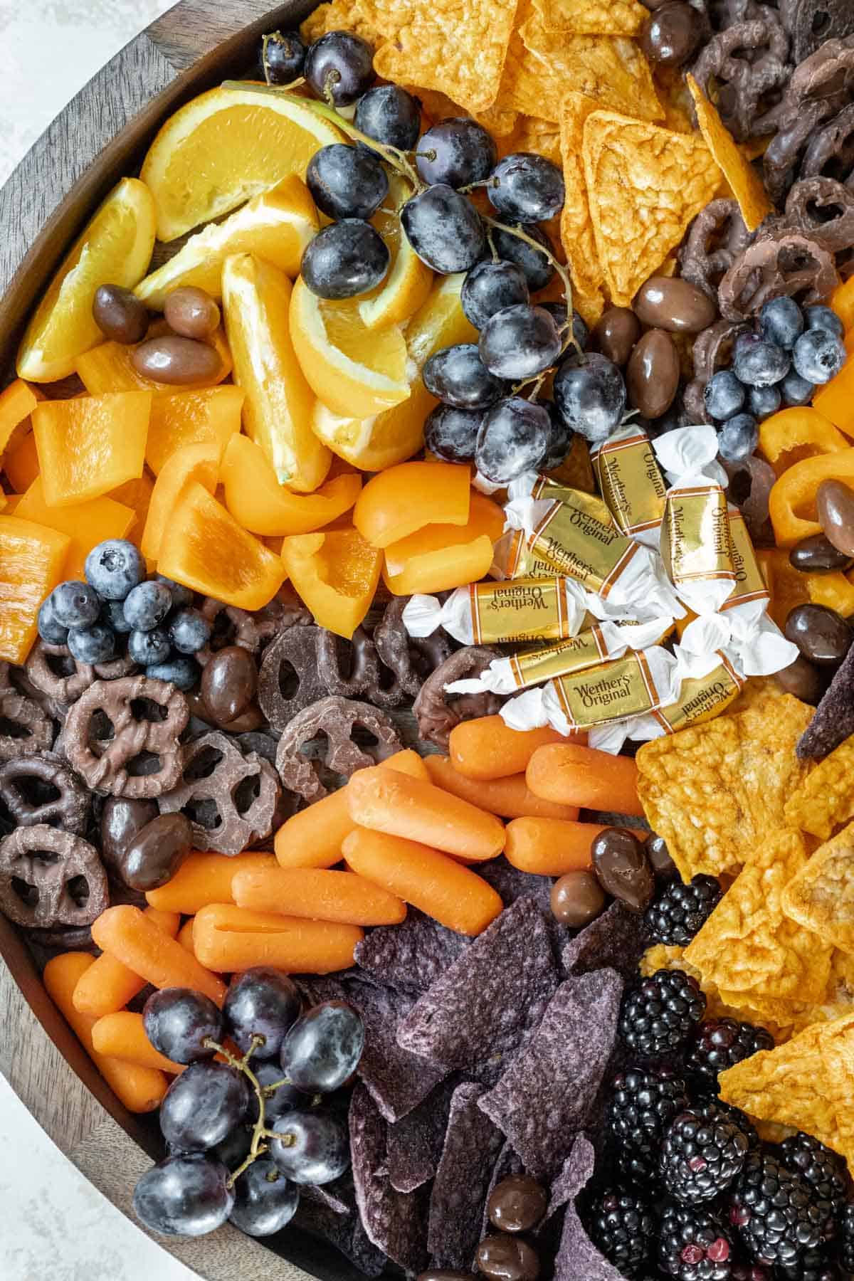 Spooky Halloween snack board with orange slices, caramels, carrots, nacho cheese chips, blue tortilla chips, chocolate pretzels, grapes, and blueberries.